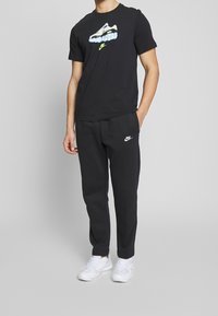 Svart bomulls-T-shirt med sneakersgrafik och logotyp, kombinerad med svarta sweatpants med vit logotyp och vita sportskor.