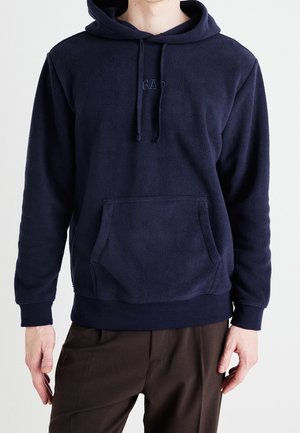 Mand iført marineblå fleece-hoodie med frontlomme og brune bukser, stående mod en ensartet hvid baggrund.