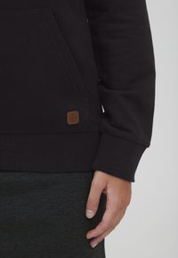 Sudadera negra con puños y dobladillo acanalados, que presenta un bolsillo tipo canguro y un parche de logo de cuero marrón en el lateral. Textura suave.