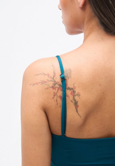 Τιρκουάζ φόρεμα με λεπτές τιράντες, που αναδεικνύει ένα λουλουδένιο τατουάζ σε αποχρώσεις του κόκκινου και του μαύρου στον ώμο, σε απλό φόντο.