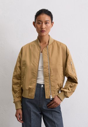 Veste bomber kaki avec une fermeture éclair à l'avant, des poignets côtelés et une poche latérale. Le tissu semble lisse avec un léger éclat.