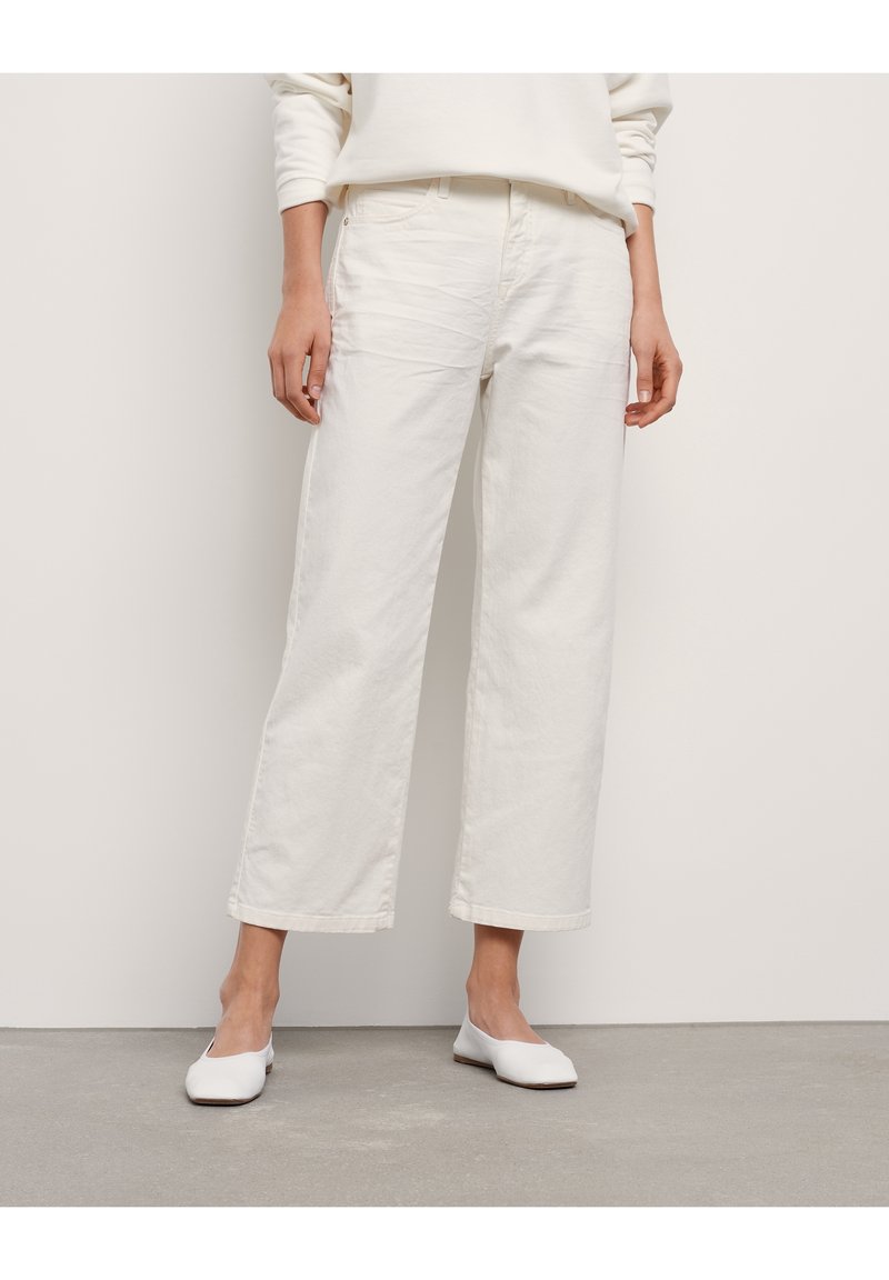 Personne portant un pantalon large court écru, un haut à manches longues écru assorti et des chaussures plates blanches, debout sur un sol gris.