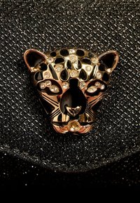 Juodas tekstūruotas krepšys su auksiniu leopardų galvos užsegimu, papuoštu juodais ir skaidriais brangakmeniais, pabrėžiančiu išskirtinį dizainą ir detales.