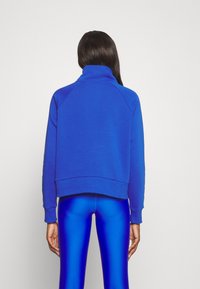 Sudadera azul eléctrico con cuello alto, mangas raglán y puños acanalados, combinada con leggins azules brillantes. Textura de tejido suave.