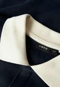 Polo navy con colletto a coste color crema, dotato di un'etichetta con il nome del brand "LMTD" e informazioni sulla taglia. Tessuto morbido e vestibilità su misura.