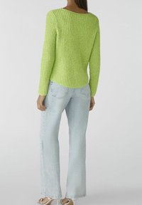 Person i en limegrøn ribbet langærmet sweater, lys blå højttaljede vide jeans og hvide hæle sandaler, står med ryggen synlig.