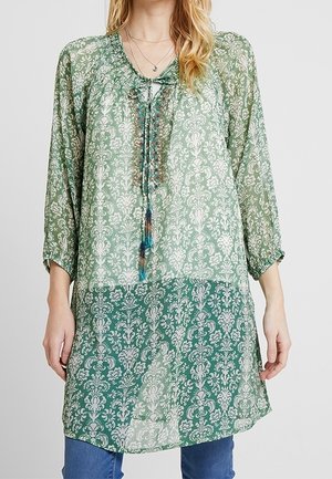 Groene tuniek met patroon, v-hals, driekwart mouwen en een strikdetail. De stof is doorschijnend met bloem- en damaskontwerpen.