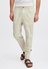 Helle beige Hosen aus einem strukturierten Stoff, mit vorderen Falten, einem Knopfverschluss und einem elastischen Bund. Getragen mit braunen Sandalen.