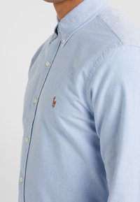 Ljusblå skjorta med knappar i bomull, med klassisk krage, långa ärmar och en liten broderad logotyp på bröstet.