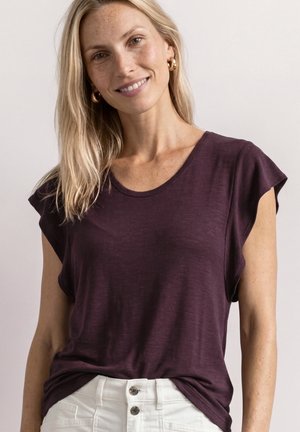 Femme blonde souriante portant des boucles d'oreilles anneaux dorés, un t-shirt violet foncé à manches courtes, et un pantalon blanc taille haute avec des boutons.
