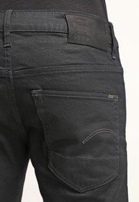 Jeans de mezclilla negra con una textura suave, que cuentan con una etiqueta de cuero, costuras mínimas y un diseño distintivo en el bolsillo trasero.
