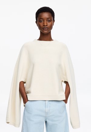 Mujer vistiendo un suéter de punto crema extragrande con mangas extralargas y jeans azul claro de talle alto, de pie con las manos en los bolsillos.