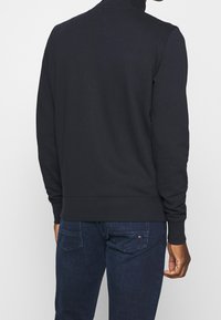 Tmavě modrý pulovrový svetr s vysokým límcem, dlouhými rukávy a žebrovanými manžetami. V kombinaci s tmavými džínami. Hladká textura a přiléhavý střih.