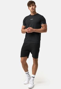T-shirt noir à manches courtes avec un graphisme contrastant sur les manches, associé à un short noir. Les chaussures sont des baskets blanches avec des accents noirs.