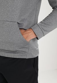 Pull-over sportif gris en tissu texturé, avec des poignets ajustés et une poche avant. Associé à un pantalon de sport foncé pour un look sportif.