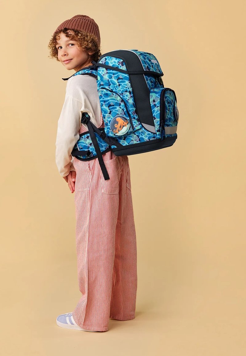 Sac à dos bleu à motifs avec des accents noirs, plusieurs compartiments et un badge rond présentant un design de pieuvre. Tissu texturé.