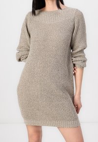 Femme portant une robe pull en tricot beige à manches longues avec un col large et des manches retroussées, sur fond uni.