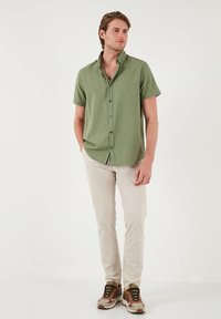 Cămașă scurtă verde deschis cu guler butonat și nasturi albaștri, asortată cu pantaloni bej și adidași multicolori.