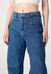 Modré denimové džíny s vyšívanými červenými růžemi a zelenými listy po stranách předních kapes, v pase na vysoké úrovni a s klasickým pětikapsovým designem.