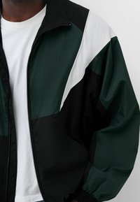 Vindjacka med svarta, mörkgröna och vita paneler, dragkedjestängning, ribbad manschett och avslappnad passform. Texturen är mjuk och lätt.