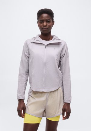 Γυναίκα με φωτεινή μωβ φούτερ με φερμουάρ, μπεζ σορτς και κίτρινα ποδηλατικά σορτς σε απλό λευκό φόντο.