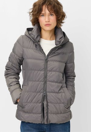 Woman with short curly brown hair wearing a gray quilted puffer jacket with a high collar and hands in pockets.