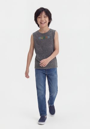 Garçon souriant marchant vers l'avant, portant un débardeur gris "GUESS", un jean bleu et des chaussures d'intérieur bleu marine sur fond blanc.