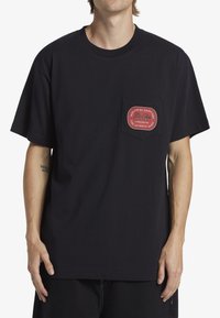 Svart bomulls-t-shirt med kort ärm, har en framficka prydd med ett röd- och vitt cirkulärt logomärke. Enkel och avslappnad passform.