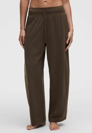 Femme portant un pantalon ample et large de couleur vert olive avec une taille élastique et un cordon, debout pieds nus devant un fond uni.