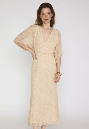 Femme portant une robe midi rose pâle à fleurs avec manches à volants, décolleté en V profond, et bracelets dorés au poignet droit, debout devant un fond uni.