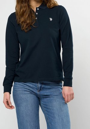 Frau trägt ein schwarzes Polohemd mit langen Ärmeln und kleinem weißem Logo sowie blaue Jeans, steht vor einem einfarbigen Hintergrund.