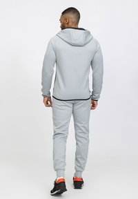 Survêtement de sport gris avec une capuche zippée, des poignets élastiques et un pantalon. Présente des accents noirs et des baskets noires avec des détails orange.