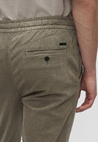 Primo piano di una persona che indossa pantaloni beige in velluto a coste con elastico in vita e tasca posteriore con bottone, braccio appoggiato accanto al corpo.