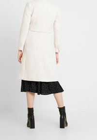 Abrigo de mezcla de lana color beige claro con un diseño ajustado, mangas largas, detalle de cinturón en la parte posterior y acentos de botones, usado sobre un vestido negro con lunares.
