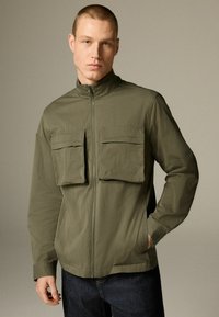 Mann in olivgrüner Zip-Jacke mit zwei großen Brusttaschen und dunkelblauen Jeans, der vor einem neutralen Hintergrund steht.