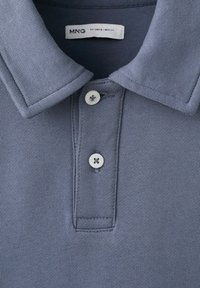 Gros plan sur le col d'un polo bleu avec deux boutons blancs et une étiquette de taille visible marquée S.