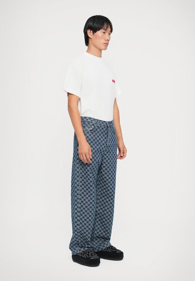 Un tricou alb cu un logo mic roșu, asortat cu blugi largi, cu un model albastru, având un design în grilă, și pantofi negri tip slip-on.