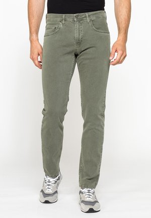 Uomo con jeans slim fit verde oliva e sneakers grigie bianche in piedi su sfondo bianco.