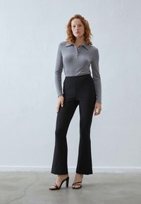 Grijze geribbelde langemouw polo, zwarte strakke flare broek, zwarte sandalen met bandjes, staand op een betonnen vloer tegen een eenvoudige achtergrond.
