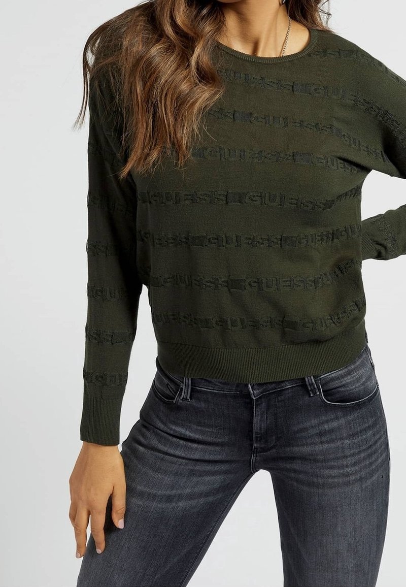 Suéter verde oliva con textura acanalada y franjas horizontales con logo. Cuello redondo y mangas largas, combinado con jeans oscuros de mezclilla.