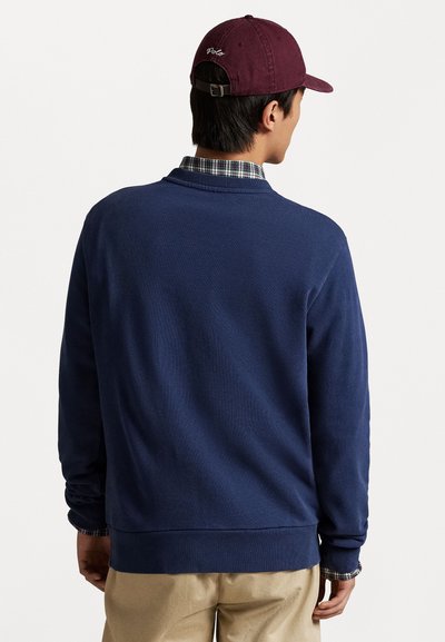 Homem de costas, vestindo um suéter azul-marinho sobre uma camisa xadrez, calças bege e um boné de beisebol bordô com o texto "Polo".