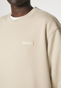 Homme portant un sweat-shirt beige Columbia par-dessus une chemise blanche, montrant la zone de la poitrine et le patch logo sur le côté gauche.