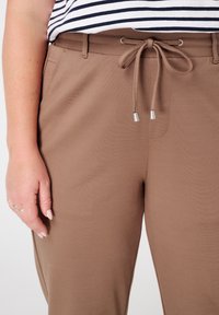 Pantalons marron à taille élastique avec détail de cordon de serrage, présentant une texture lisse et une coupe ajustée, associés à une chemise rayée au-dessus.