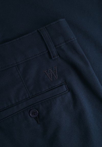 Pantalon bleu marine en tissu texturé, avec un bouton visible et un subtil logo "W" brodé sur la poche.