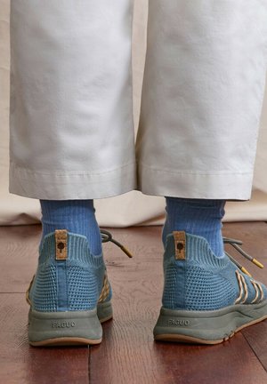 Pieds chaussés de baskets en maille bleue et de chaussettes bleues, debout sur un sol en bois, avec un pantalon blanc raccourci visible de derrière.
