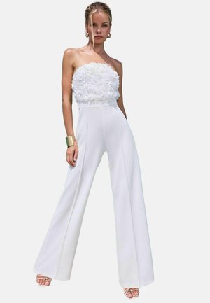 Hvid jumpsuit med en strapless, blomsterstruktureret overdel, brede ben og et glat stof. Accenteret med guldsmykker og sandaler med remme.