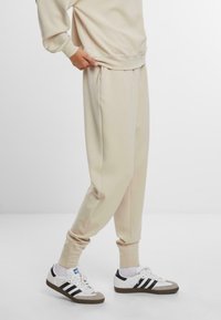 Beige joggebukser i bløt stoff med elastiske mansjetter og sidelommer, kombinert med hvite sneakers med svarte striper og grå detaljer.