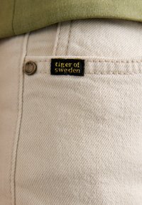 Vit denimtyg med en liten svart etikett med "tiger of sweden" i gul text, fäst på en ficka. Enkel struktur och sömmar synliga.