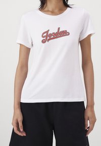 Bílý bavlněný tričko s kulatým výstřihem a krátkými rukávy. Na hrudi je červené logo "Jordan" v zakřiveném, stylizovaném písmu.