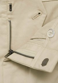 Tissu beige en gros plan montrant une fermeture éclair métallique partiellement ouverte, une boutonnière, un bouton transparent et un petit détail en métal en forme de crâne sur un poignet.